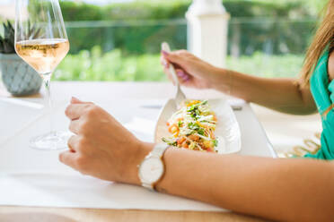 Side view of unrecognizable female sitting at table with plate of appetizing salad made of vegetables and shrimps garnished with grated cheese while having lunch in patio of restaurant in summer - ADSF46990