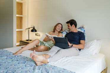 Lächelnde Freundin, die ein Buch auf dem Bett liest, neben einem fröhlichen Freund, der auf einem Tablet surft, während sie das Wochenende zusammen im Schlafzimmer verbringen - ADSF46991