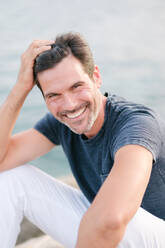 Delighted adult bearded male tourist in white pants and t shirt leaning on hand and smiling at camera while sitting on rocky coast near sea on sunny day - ADSF46992
