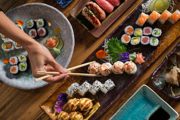 Von oben der Ernte anonyme Person mit Stäbchen essen verschiedene Sushi-Rollen auf dem Tablett an Holztisch im Café serviert - ADSF47005