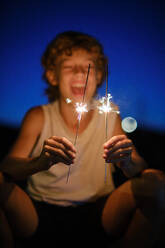 Excited preteen boy looking at burning sparklers and enjoying celebration of holiday on background of night sky - ADSF47007