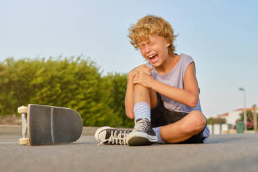 Ground level of unhappy preteen skater embracing injured knee and suffering from pain after falling off skateboard while sitting on road and shouting with closed eyes - ADSF47011