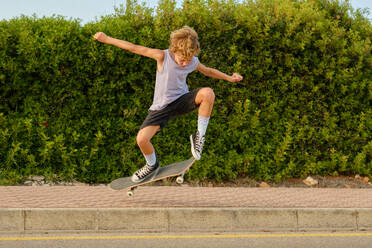 Aktiver Teenager, der mit dem Skateboard über eine asphaltierte Straße springt und einen Trick zeigt, während er im Sommer Spaß hat - ADSF47013