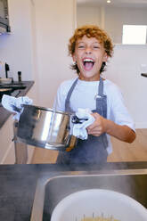 Happy child in apron with pot looking at camera against sink with pasta in colander at home - ADSF47015