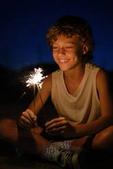 Fröhlicher Junge im Teenageralter, der mit glühenden Wunderkerzen in der Nacht sitzt und einen festlichen Anlass feiert - ADSF47017