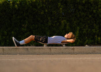 Ebenerdige Seitenansicht eines Teenagers, der auf einem Skateboard auf der Straße liegt und den Sonnenuntergang im Sommer genießt - ADSF47020