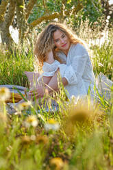 Sensible junge Frau in weißem Sommerkleid, die ihr gewelltes Haar berührt und in die Kamera schaut, während sie auf einer Wiese in einer sonnigen Sommerlandschaft sitzt - ADSF47023