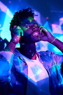 Zufriedener afroamerikanischer männlicher DJ in legerer Kleidung, der über Kopfhörer Songs hört, während er mit geschlossenen Augen in einem dunklen Studio mit leuchtenden Neonröhren steht - ADSF47030