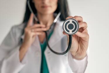 Crop unrecognizable female physician in white medical robe with stethoscope during checkup on light background - ADSF47034