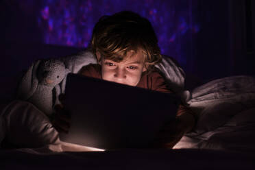 Cute boy in sleepwear lying on bed under blanket while browsing tablet in dark bedroom at night time in flat - ADSF47039