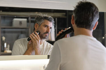 Konzentrierter Mann in Freizeitkleidung rasiert seinen Bart mit einem professionellen Trimmer, während er sein Spiegelbild zu Hause betrachtet - ADSF47043