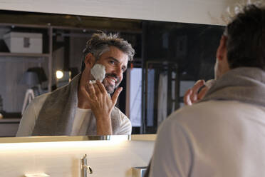 Smiling man standing in modern bathroom near mirror and looking at reflection while applying shaving foam and doing morning skin care routine at home - ADSF47044