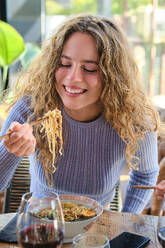 Positive junge Frau sitzt an einem gedeckten Tisch und isst würzige Ramen mit Stäbchen in einem modernen Restaurant am Wochenende - ADSF47059