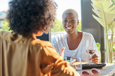 Fröhliche afroamerikanische Frau, die lächelt und sich gegenseitig anschaut, während sie mit einem Glas Rotwein in einem Restaurant am Tisch sitzt - ADSF47067