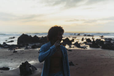 Young man holding sunglasses standing at beach - PBTF00254