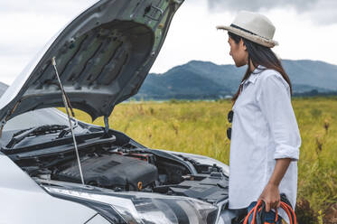 Asian woman holding battery booster cable copper wire for repairing breakdown broken car by connect with red and black line to electric terminal by herself. Car maintenance and transportation concept - INGF12084