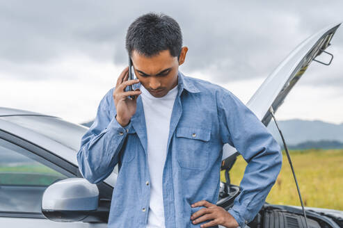 Asian man calling car maintenance service after having accident during travel long weekend between road trip. Car broken in mountain meadow background. People transportation. Need help insurance claim - INGF12085