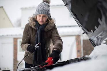 Woman Using Jumper Cables On Car Battery On Snowy Day - INGF12096