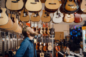 Bearded young guy choosing acoustic guitar in music store. Assortment in musical instruments shop, male musician buying equipment. Young guy choosing acoustic guitar in music store - INGF12101