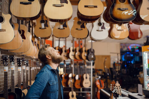 Bearded young guy choosing acoustic guitar in music store. Assortment in musical instruments shop, male musician buying equipment. Young guy choosing acoustic guitar in music store - INGF12101