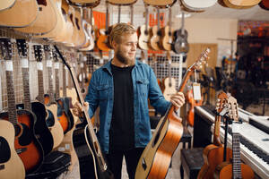 Bearded young guy choosing acoustic guitar in music store. Assortment in musical instruments shop, male musician buying equipment. Young guy choosing acoustic guitar in music store - INGF12102