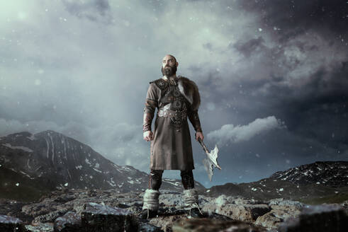 Viking with axe dressed in traditional nordic clothes standing in rocky mountains. Scandinavian ancient warrior - INGF12103