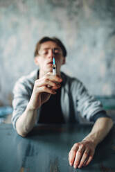 Male junkie sitting at the table with syringe in hand, grunge room interior on background. Drug addiction concept, addicted people. Junkie sitting at the table with syringe in hand - INGF12113