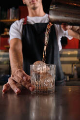 Gesichtsloser männlicher Barkeeper in schwarzer Schürze, der am Tisch steht und mit einem Shaker einen Cocktail mixt, während er in einer Bar arbeitet - ADSF47072