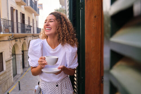 Positive young female with eyes closed laughing while drinking tea and standing on balcony over narrow city street - ADSF47076