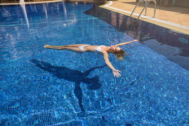 Full length of carefree young woman in white bikini swimming in clean pool with eyes closed and spreading arms while resting in tropical resort during summer vacation - ADSF47077