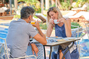 Fröhliche Frau, die eine Tasse Kaffee trinkt, während sie Zeit mit einem Mann verbringt, der an einem Tisch in einem Resort in der Nähe des Pools an einem sonnigen Tag Saft trinkt - ADSF47081