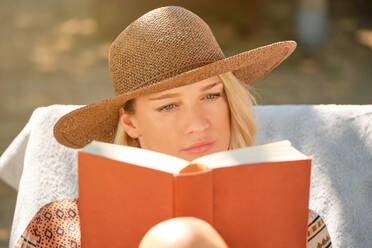 Pensive young female in straw hat and jacket looking at red bounded book while reading with concentration and sitting on white mattress on deckchair - ADSF47089