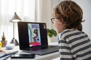 Side view of cute kid having video call with classmate via laptop while sitting at table with smartphone in light room - ADSF47101