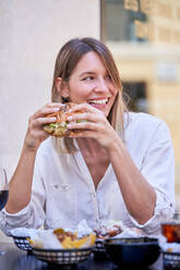 Positive junge Frau mit blonden Haaren, die an einem Tisch mit Essen und Getränken sitzt und einen appetitlichen Hamburger auf der Terrasse eines Cafés isst, während sie wegschaut - ADSF47105