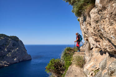 Seitenansicht eines Mannes mit Rucksack, der auf einem steinernen Fußweg auf einer Klippe steht und das plätschernde Meer im Sommerurlaub beobachtet - ADSF47109