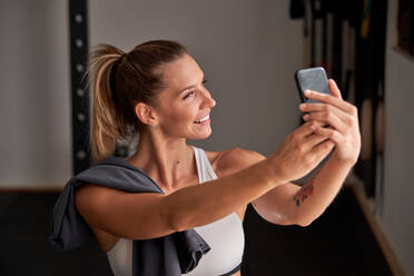 Positive young female athlete with ponytail in sports bra taking selfie on smartphone in modern gym after workout - ADSF47111