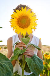 Unerkanntes Kind in ärmellosem T-Shirt, das sein Gesicht mit einer blühenden Sonnenblume bedeckt, während es auf einem unscharfen Feld an einem sonnigen Sommertag steht - ADSF47114