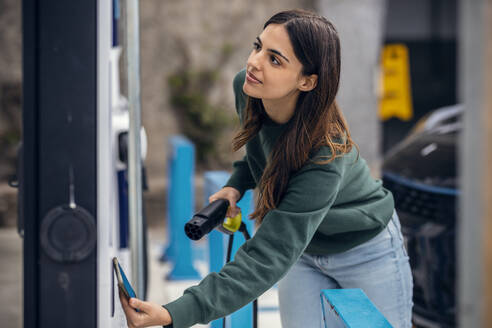 Young woman paying through smart phone at electric vehicle charging station - JSRF02609