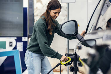 Lächelnde Frau beim Einstecken des Ladegeräts in ihr Auto an einer Ladestation für Elektrofahrzeuge - JSRF02612