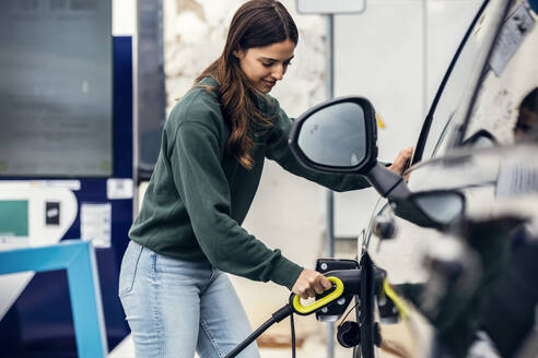 Smiling woman plugging charger in car at electric vehicle charging station - JSRF02612