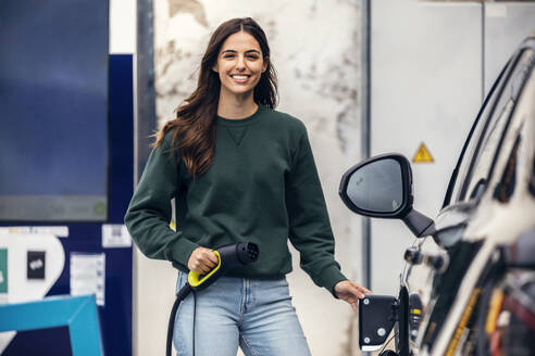 Happy young woman standing with electric plug at vehicle charging station - JSRF02613