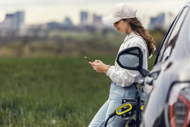 Junge Frau mit Mütze, die ihr Smartphone benutzt und ihr Auto an einer Tankstelle auflädt - JSRF02614