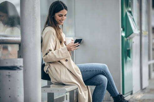 Glückliche Frau sitzt auf einer Bank und benutzt ein Smartphone - JSRF02622