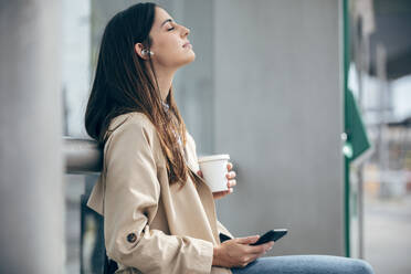 Young woman wearing wireless in-ear headphones listening to music - JSRF02624