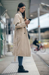 Lächelnde Frau mit Smartphone an der Straßenbahnhaltestelle - JSRF02625