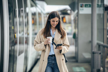 Glückliche Frau mit Kaffeetasse und Smartphone an der Straßenbahnhaltestelle - JSRF02626