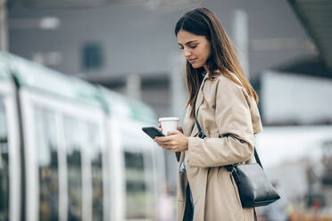 Junge Frau mit Kaffeetasse und Smartphone am Bahnhof - JSRF02628