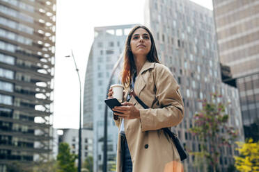 Thoughtful young woman standing with smart phone and coffee cup - JSRF02631