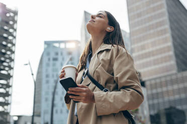 Young woman with eyes closed holding coffee cup and smart phone in city - JSRF02632