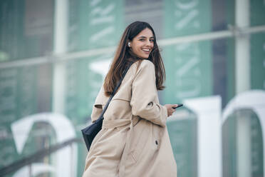 Happy young woman holding smart phone in front of building - JSRF02646